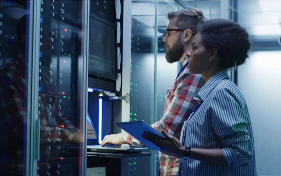 Two business people standing and working together with a laptop and clipboard as they monitor a data server in glass cases.