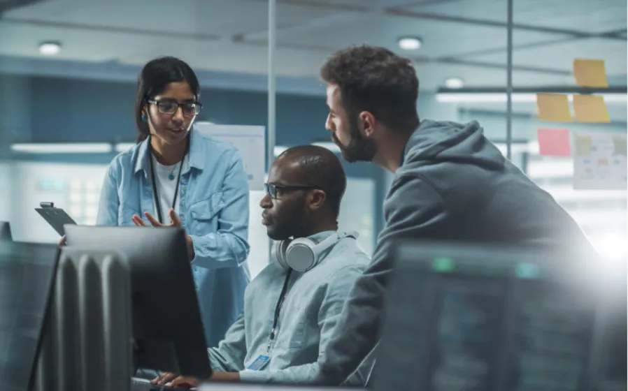 A man with headphones sitting at a computer monitor in an office with colleagues standing next to him conversing.