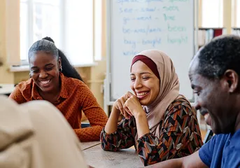 Group of diverse employees sitting at a table laughing during a meeting with a whiteboard in the background.