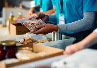 Close up of volunteer wearing blue shirt packing food into plastic bins at a food bank.