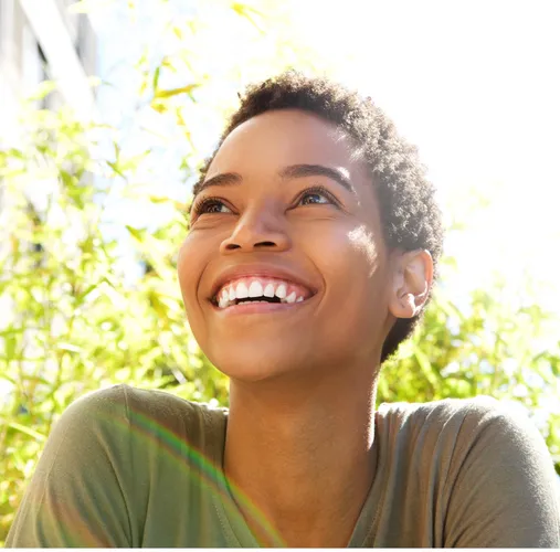 A smiling young woman looking off into the distance while sitting outside in front of a tree on a bright and sunny day.