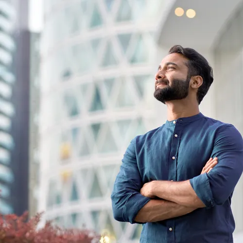 A man standing, crossing his arms, and smiling in a city during the day as he looks off to the side.