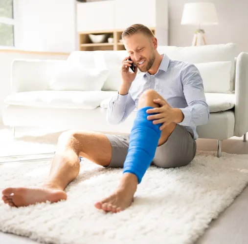 A man with a leg bandage sitting on the floor of a residential room in front of a couch while he talks on his smartphone.