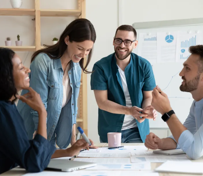 Four smiling and laughing colleagues brainstorming a financial concept while looking at paper printouts at an office desk. 