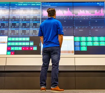 A man with his back to the camera examining a big display of screens showing bar graphs and detailed reports.