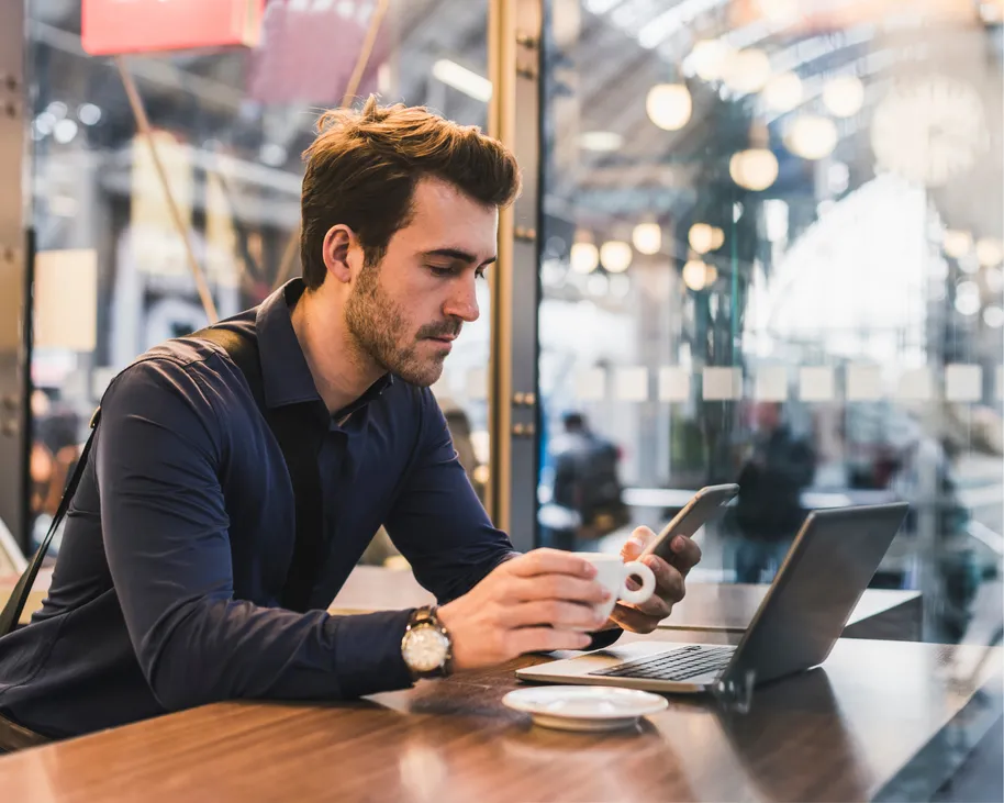 Professional man sitting at a café while he holds a coffee cup and looks at his smartphone.