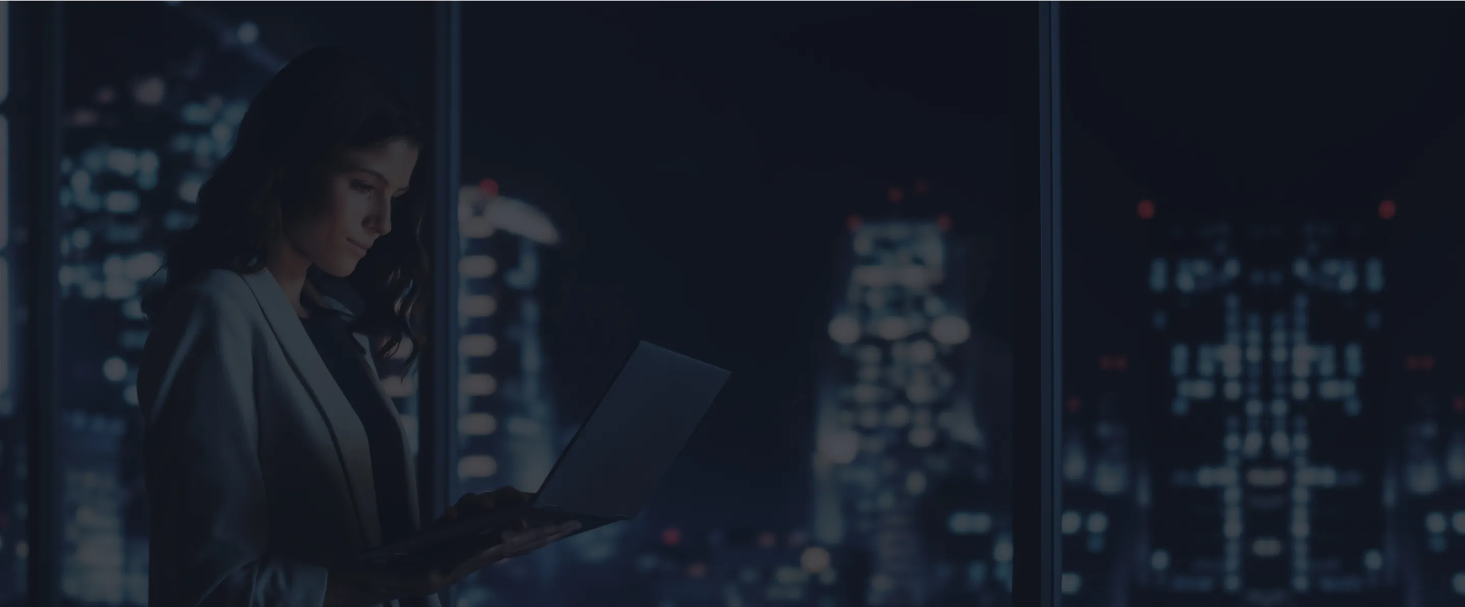 A woman working on a laptop in an office with a nighttime skyline view.