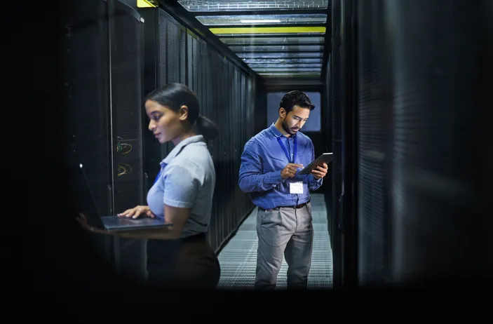 Two professionals monitoring database safety in a data server center by using software on a laptop and a smartphone.