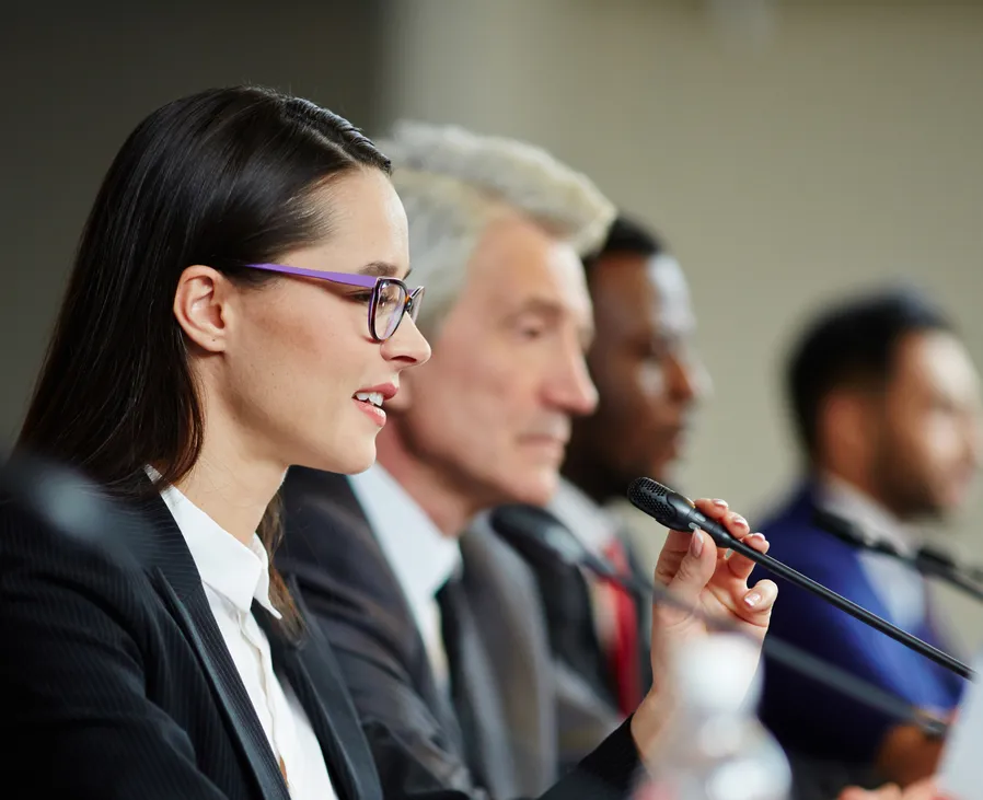 Row of professional speakers wearing suits and sitting in front of microphones to answer questions from an audience.